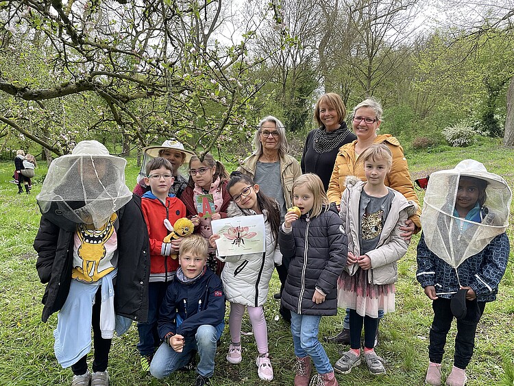Kindergruppe, Kinder teils mit Imkerhüten, drei Frauen, blühender Apfelbaum.